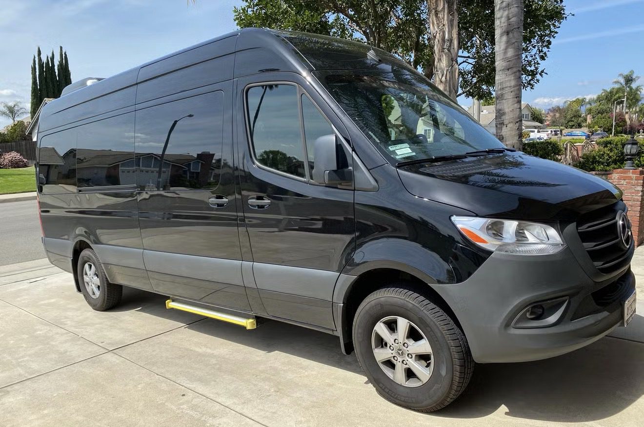 A black cargo van parked on a driveway beside trees.
